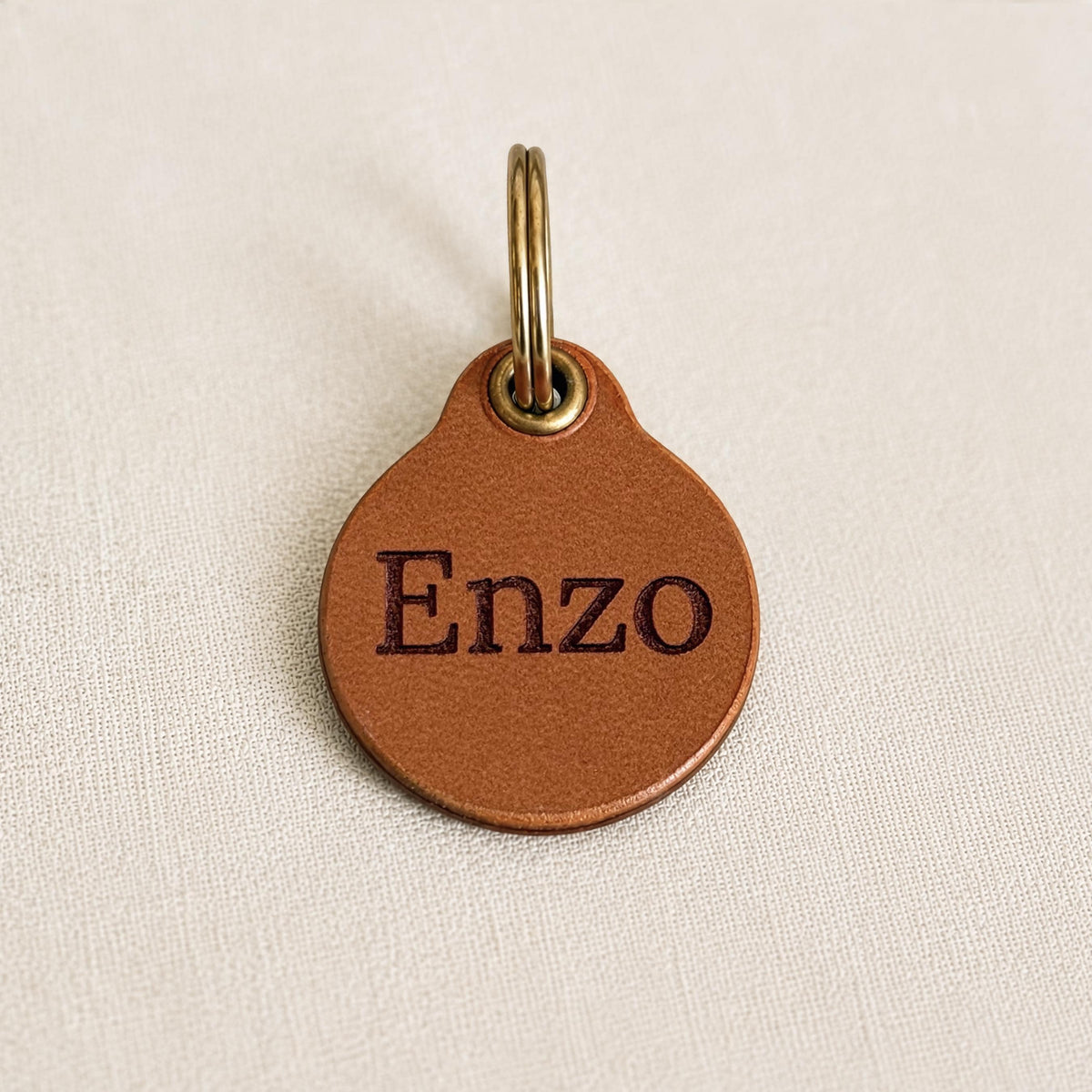May include: A round, tan leather tag featuring the name Enzo embossed in dark brown lettering. The tag includes a brass-coloured metal eyelet at the top, which is attached to a circular brass-coloured metal split ring for secure attachment.