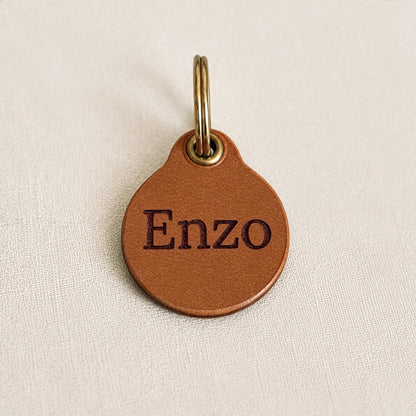 May include: A round, tan leather tag featuring the name Enzo embossed in dark brown lettering. The tag includes a brass-coloured metal eyelet at the top, which is attached to a circular brass-coloured metal split ring for secure attachment.