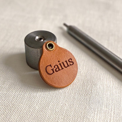 May include: A round tan leather tag featuring the embossed name Gaius, a brass-coloured metal eyelet at the top, a cylindrical metal tool base, and a long, thin, silver-coloured metal rod positioned diagonally across the frame.